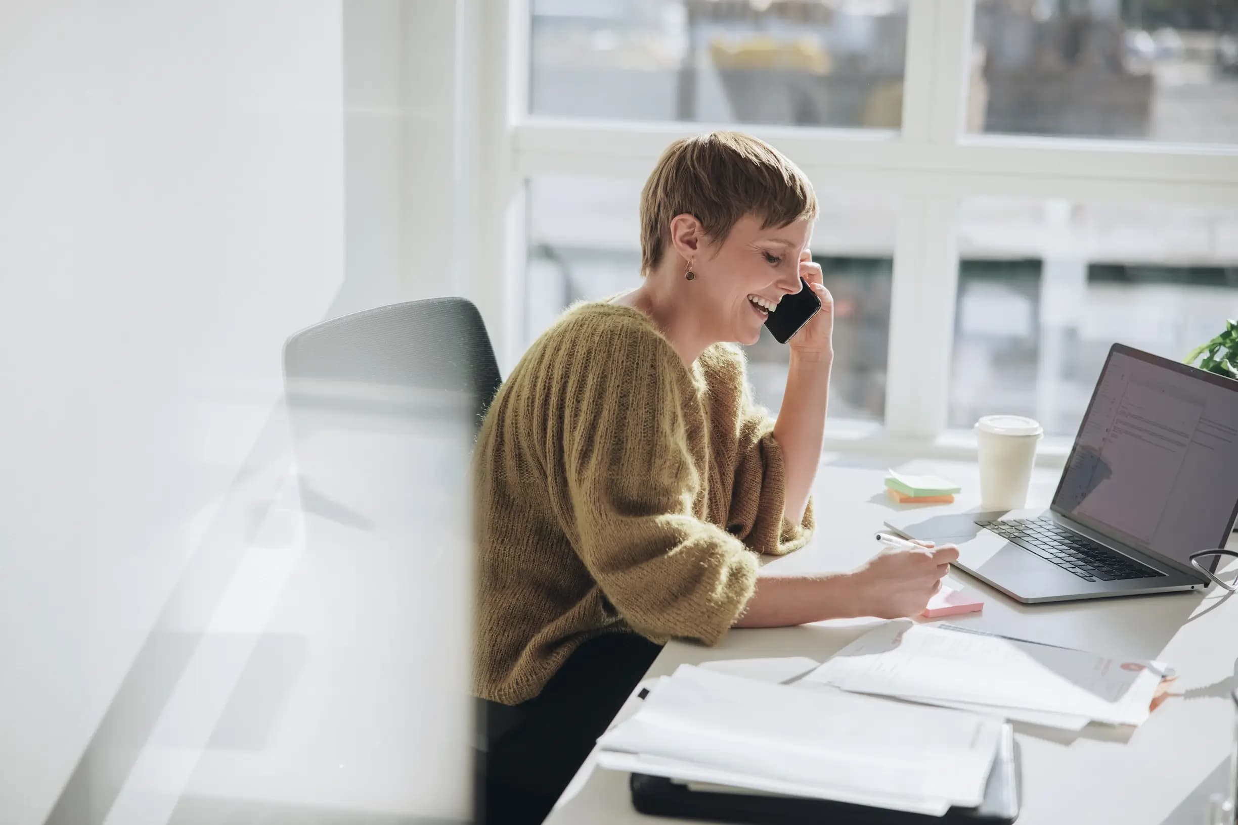 Mitarbeiterin telefoniert am Schreibtisch und arbeitet mit Notebook und Stift und Zettel Mitarbeiterin telefoniert am Schreibtisch und arbeitet mit Notebook und Stift und Zettel