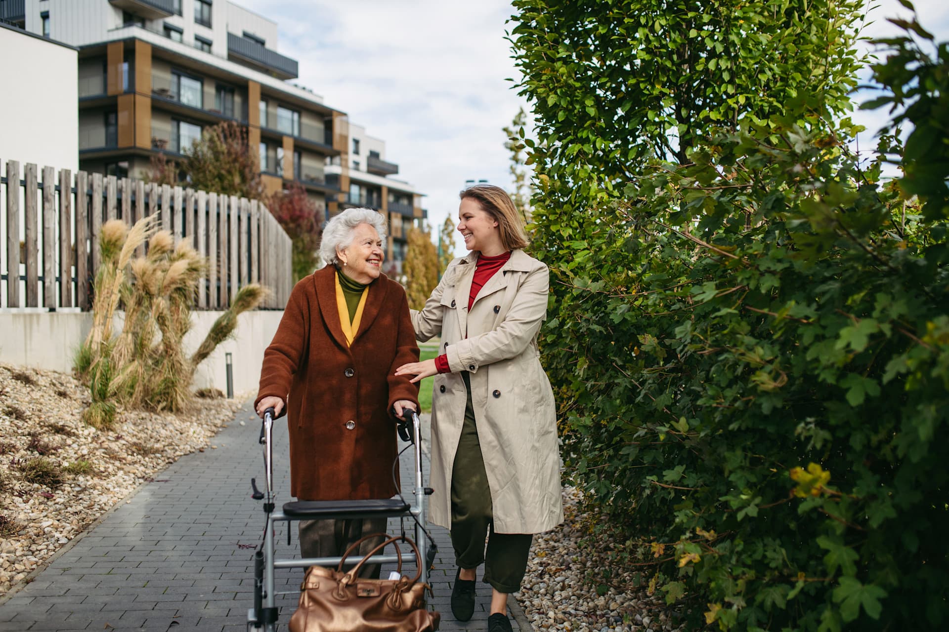 In einer Wohnsiedlung spazieren eine Seniorin mit Rollator und eine jüngere Frau nebeneinander In einer Wohnsiedlung spazieren eine Seniorin mit Rollator und eine jüngere Frau nebeneinander