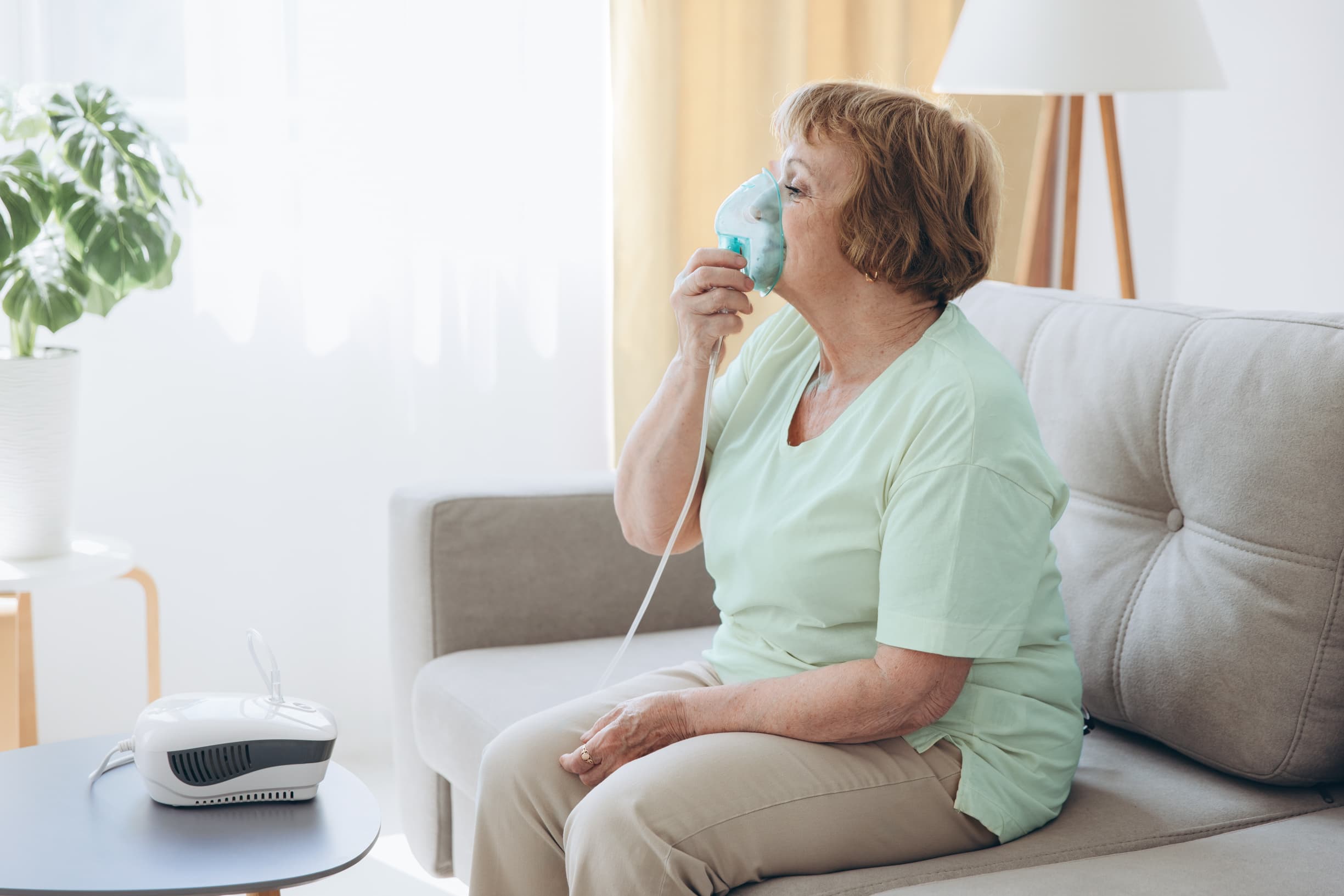 Eine Seniorin, die aufrecht auf dem Sofa sitzt und sich eine Beatmungsmaske vor ihren Mund und ihre Nase hält Eine Seniorin, die aufrecht auf dem Sofa sitzt und sich eine Beatmungsmaske vor ihren Mund und ihre Nase hält