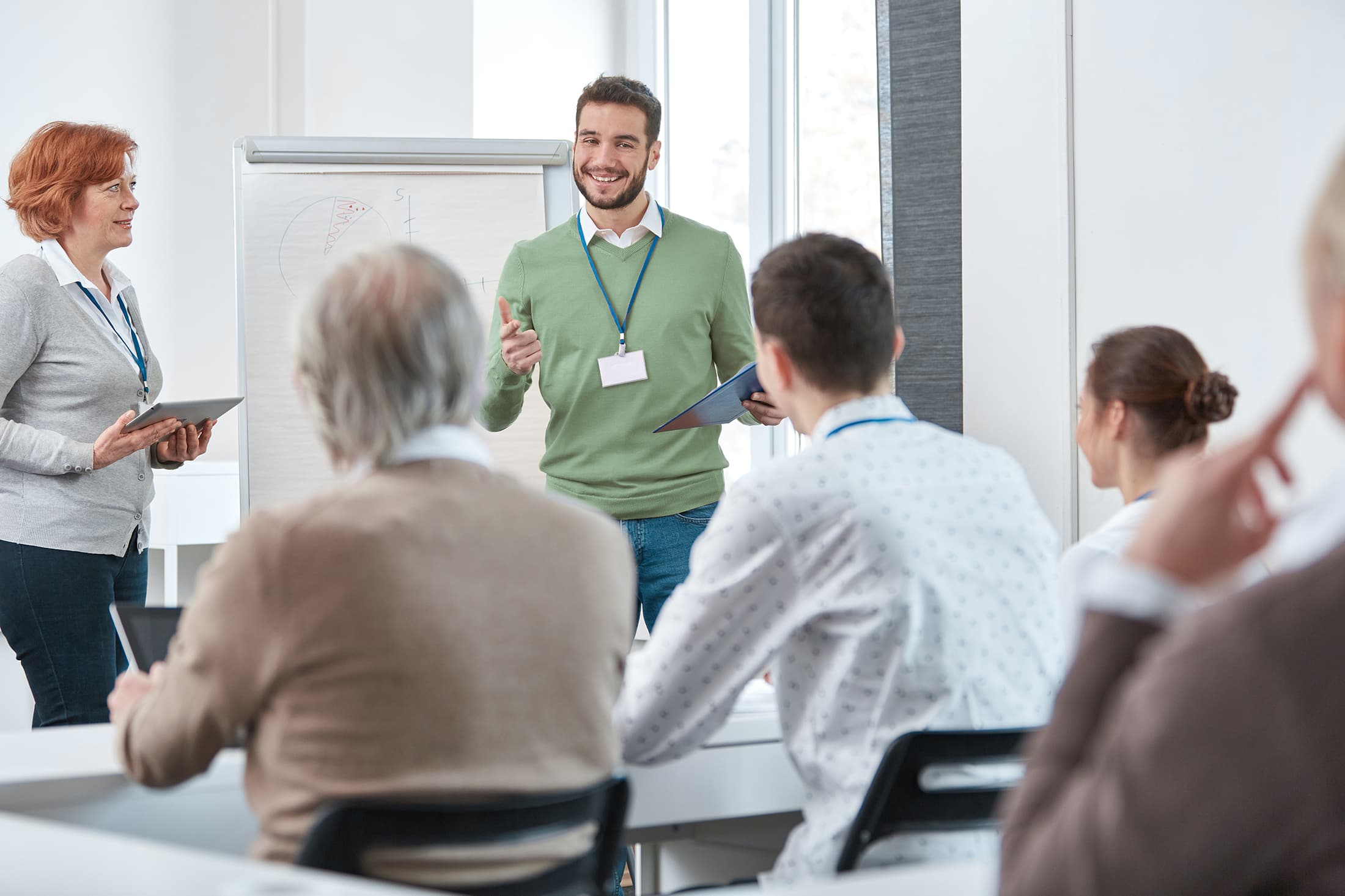 Mann, der in einem Schulungsraum als Dozent vor einer Teilnehmergruppe steht Mann, der in einem Schulungsraum als Dozent vor einer Teilnehmergruppe steht