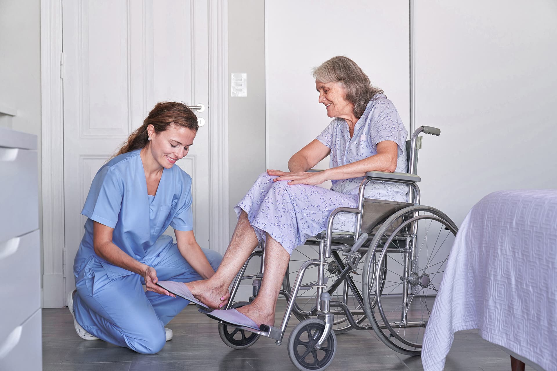 Eine Pflegekraft kniet vor einer Patientin, die im Rollstuhl sitzt und hilft ihr dabei, ihre Schlappen anzuziehen Eine Pflegekraft kniet vor einer Patientin, die im Rollstuhl sitzt und hilft ihr dabei, ihre Schlappen anzuziehen